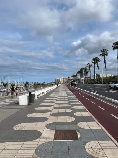 The image shows a view of the promenade at Las Palmas De Gran Canaria, Spain. The perspective is along a pathway paved with gray and white patterned tiles, with a distinct design featuring semicircular shapes. Adjacent to the pathway is a red bike lane.
Along the upper edge of the image is a road with palm trees lining the side, and a white car is parked.
On the left side, there's a view of a marina filled with boats. Two people are walking near a barrier along the marina. The sky is partly cloudy with patches of blue.