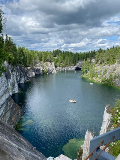 This is a vertical, high-angle photograph of Ruskeala Mountain Park in Russia. The scene depicts a quarry filled with clear, dark blue water, surrounded by steep, rocky cliffs and dense green forests. The sun is not directly visible, but the bright sky with dramatic, grey and white clouds suggests it is daytime, likely late morning or afternoon, and the weather is mixed with sun and clouds.
Several small boats, likely kayaks or canoes, are visible on the water, with people wearing orange life vests. One boat is in the middle distance, and another is closer to the shore on the left. There are also some small yellow buoys forming a line in the water.
The rocky cliffs display variations in texture and color, with some areas appearing smooth and others rough and fragmented. Green vegetation, including pine trees, grows abundantly on the cliffs and along the top edges. In the upper right corner, a metal railing and some white wildflowers are visible, indicating the vantage point is from a bridge or elevated walkway. The overall mood is serene and picturesque, highlighting the natural beauty of the location. No visible text is present in the image.