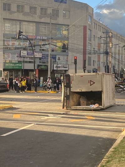 This is a daytime, eye-level, street-level photograph taken in Quito, Ecuador, likely in the late afternoon given the long shadows. The scene depicts a busy urban environment with a multi-story building on the left, featuring numerous windows and advertisements for dental services, including "CENTRO DE FRESADO" and "NATURAL VITAE". A traffic light shows a red signal.
In the foreground, a white box truck is parked on the street, its rear doors open. A group of people, many appearing to be young adults, are gathered around the truck and on the sidewalk. Some are standing, while others are seated or leaning. Their expressions are neutral, and there is no overt indication of distress or celebration. The activity appears to be a casual gathering or waiting period, possibly related to the contents of the truck or simply a meeting point.
The truck has "ECUACERD" and a license plate "PBY-4309" visible, along with a triangular logo. A motorcycle is parked further down the street. The presence of overhead electrical wires and a sidewalk with grass and pavement indicates a typical urban setting. The overall impression is that of an ordinary day in a city, with people congregating on the street. Visible text includes company names and a phone number on the building, as well as text on the truck.