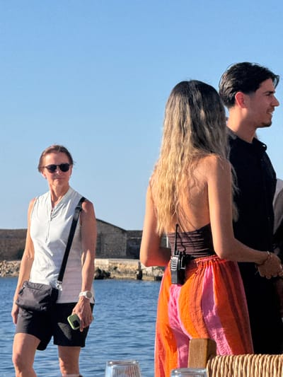 In Chania, Greece, a group of three individuals stands by the sea under a clear blue sky. On the left, a woman with short brown hair and sunglasses wears a white sleeveless top and black shorts, with a black crossbody bag and a green smartphone in her right hand. To her right, a young woman with long blonde hair, seen from behind, is wearing a dark top and a brightly colored striped skirt. A black walkie-talkie is clipped to her waistband. Next to her, a man with dark hair, also seen from the side, wears a black shirt. In the background, a low stone building and the blue sea are visible.