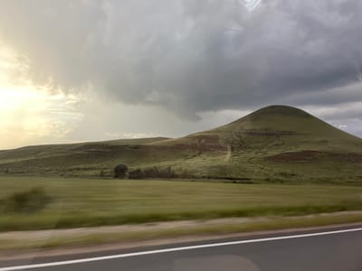 The image depicts a landscape under a dramatic, cloudy sky, likely taken from a moving vehicle due to motion blur in the foreground and reflection on the upper portion of the frame. In the middle ground, rolling green hills rise, with a distinctly conical peak dominating the right side of the image. The hills are covered in short, green vegetation, with some reddish-brown streaks suggesting exposed soil or rock formations. A strip of dark trees is visible along the base of the hills. In the foreground, a blurred expanse of green fields stretches across the scene, bordering a dark asphalt road with a white line. The sky is a mix of bright, sunlit clouds on the left, transitioning to darker, heavier storm clouds on the right, creating a sense of changing weather. The location is specified as Kipchak-Askarovo, Russia. There are no people or animals visible in the image.