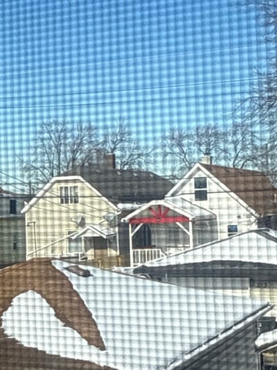 Two houses with gabled roofs stand side by side under a clear, blue sky. The houses feature white trim, including railings and stairs, with one displaying a red, wheel-like structure. Bare tree branches are visible in the background. A portion of a larger building, possibly a barn or larger house, is visible on the right, its roof covered with a white material, possibly snow.