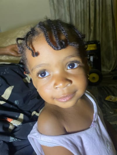 A young Black girl with braided hair and bright eyes looks at the camera. She has a neutral expression with a slight smile. She is wearing a light-colored tank top with lace trim and a gold earring. The background includes patterned bedding and a glimpse of a machine or equipment with a yellow wheel. The image is taken from a close-up angle.