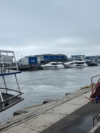 The image depicts a waterfront scene at Sunseeker International's shipyard in Poole, United Kingdom. In the foreground, the stern of a ferry or boat is visible on the left, with its white hull and railing. The water in the harbor is choppy, reflecting the overcast sky. A wooden and concrete embankment runs along the right side of the image, with a yellow rope tied to a post. Several large, modern white yachts are moored in the middle ground, docked alongside industrial buildings with blue roofs. One of these buildings features a prominent blue sign with the "Sunseeker" logo. In the background, more industrial structures and cranes are visible, indicating a working shipyard. A red metal structure and a blue cleaning bottle are present on the embankment in the lower right corner. The overall atmosphere is industrial and maritime.