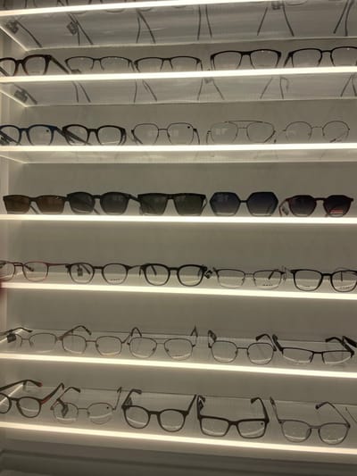 The image displays an indoor retail optical display system in Kfar Saba, Israel. Seven white, horizontally illuminated shelves are stacked vertically, showcasing numerous pairs of eyeglasses and sunglasses. The shelves are evenly spaced against a light-colored background.
The top two shelves, and shelves four through seven, primarily feature prescription eyeglasses. These include full-rim, half-rim, and rimless designs, crafted from metallic and plastic materials. Frame shapes vary, encompassing rectangular, round, and aviator styles. The third shelf from the top is dedicated entirely to sunglasses, characterized by dark-tinted lenses set in frames of diverse shapes, including rectangular, square, oversized hexagonal, and classic wayfarer-like designs.
Several frames exhibit visible branding, specifically "POLAR" on some sunglasses and a distinct "V" logo on multiple eyeglasses and sunglasses. Small informational or price tags are attached to many of the displayed items. No individuals are present in the scene, and no actions or interactions are depicted; the eyewear is statically arranged for retail presentation.