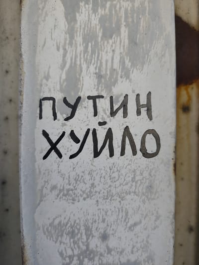 The image shows a close-up view of a weathered, light-colored metal surface with dark graffiti. The graffiti is written in Cyrillic script and appears to be a political slogan. The text is located centrally on the image and is the primary subject. The metal surface is textured and appears to be part of an outdoor structure, possibly a fence or a column, given the visible rust on the right edge of the frame. The lighting suggests it is daytime, and the overall impression is one of urban decay or neglect. No people are visible, and the scene is static. The text reads "ПУТИН ХУЙЛО", which is a derogatory phrase often directed at Vladimir Putin. The location context provided (Police, Poland) suggests this graffiti might be found in a public space within Poland, potentially as a form of protest or expression of political sentiment.