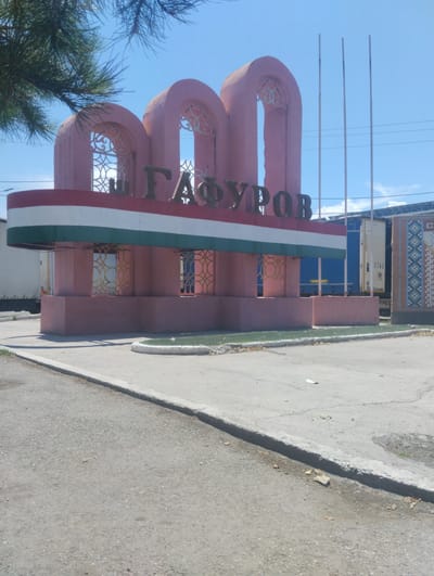 The image shows a monument in the city of Ghafurov, Tajikistan. The monument is pink and features three arched openings with intricate patterns inside. The name "ГАФУРОВ" is written in large, dark letters across the top. Below the name, there is a horizontal band of three stripes: red, white, and green. The base of the monument is also pink, with decorative elements resembling windows. In the background, there are large trucks, utility poles, and a bright blue sky. To the right of the monument, there is a section of wall with a colorful, patterned mosaic. The foreground shows a paved road and sidewalk. The overall scene depicts an outdoor public space in what appears to be a sunny day.