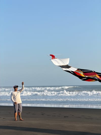 The image displays an adult male flying a large, intricate kite on a black sand beach in Selemadeg, Indonesia, under a clear, light blue sky. The man, appearing to be of Asian descent, stands barefoot on the dark sand, wearing an off-white long-sleeved shirt, dark grey shorts, a backward red and white baseball cap, and dark sunglasses. A colorful tattoo is visible on his left lower leg. His right arm is raised, holding the kite string, and he directs his gaze towards the kite positioned in the mid-right portion of the frame.

The kite consists of a prominent white, boat-like leading section with a red fin-like component at its rear. Attached to this is a long, flowing tail section composed of black, red, yellow, and orange fabrics arranged in distinct geometric patterns. The thin kite string extends from the man's right hand to the white section of the kite.

The dark sand of the beach shows subtle footprints. In the background, the ocean features multiple lines of breaking white-capped waves, extending to a dark blue-grey expanse under a pristine, cloudless sky.