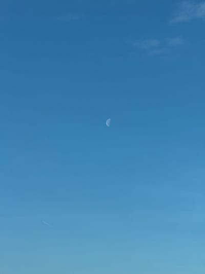A bright blue sky dominates the scene. A thin crescent moon sits slightly off-center towards the top of the frame. Faint wisps of clouds are visible in the upper left. In the bottom right, a subtle white trail, possibly a contrail from an airplane, stretches diagonally. The sky's hue deepens slightly from left to right.