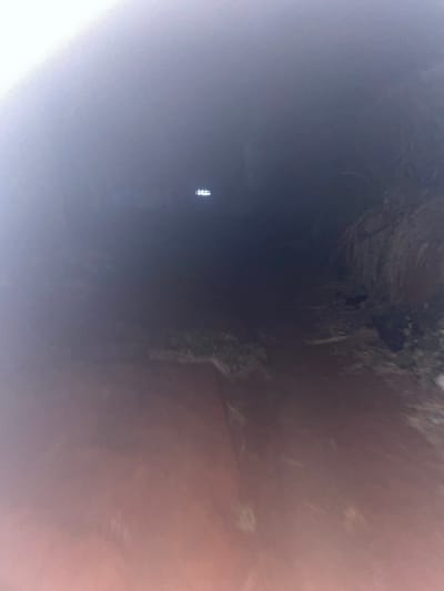 A dark, cavernous space is captured with a point of light visible in the distance. The walls are textured with rock formations and a smattering of debris. Hues of brown and grey mingle in the cavern's depths, while a hazy light seems to emanate from the left side of the scene, obscuring some of the details. The composition lacks crisp focus, presenting a slightly blurry and indistinct view of the interior space.