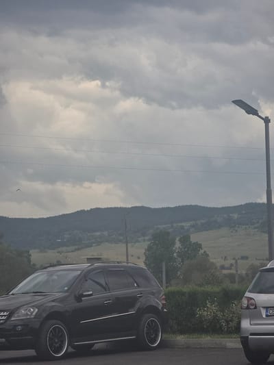 The image, when viewed upright, presents an overcast outdoor scene featuring parked cars against a backdrop of distant hills and a cloudy sky.
In the foreground, a dark-colored SUV or crossover is parked on asphalt pavement, with its front section, including the right wheel and headlight, visible. To its left, a strip of green bushes and grass separates the pavement from a partially visible silver car. The rear of the silver car, its taillight, and a Bulgarian license plate starting with "CB" are discernible.
The mid-ground reveals a landscape extending into a valley or field, bordered by several distant hills or mountains. These geographical features are densely covered with trees and appear somewhat hazy against the light. Several thin power lines traverse the upper mid-ground, originating from a tall utility pole with an attached street light on the left side of the frame. A small, dark bird is captured in flight against the cloudy sky in the central-right portion of the image.
The sky is largely covered by heavy, grey and white clouds, indicating an overcast day. The lighting is diffused and soft, without direct sunlight. The scene suggests a calm, possibly rural or semi-rural setting, likely during daytime, given the natural light despite the clouds. The "CB" on the license plate indicates a Bulgarian vehicle, consistent with the provided location context of Kraishte, Bulgaria.