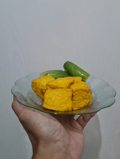 A hand holds a clear, shallow dish containing bright yellow cubes and several green cucumbers. The cubes appear to be tofu or a similar food item. The dish has a fluted edge and sits at an angle, showcasing the contents. The background is a plain, light grey wall.