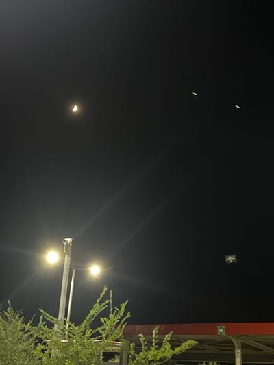 In the night sky above Unknown, Uzbekistan, a crescent moon hangs in the upper left quadrant of the frame. Below it, two streetlights with visible light flares illuminate the scene. A lamppost with a security camera is positioned in the lower center, with two bright light bulbs. In the foreground, green foliage from a tree obscures the bottom part of the image. In the middle right of the sky, a small, blurry object is visible, possibly a drone or a bird. There are also three faint, elongated green lights in the upper right of the sky.