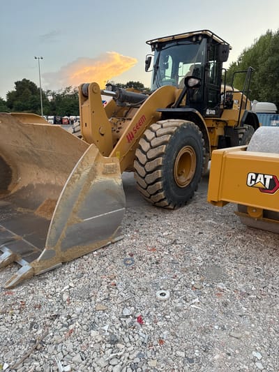 The image shows a construction site in Grenay, France, featuring heavy machinery. In the foreground, a large yellow wheel loader with a wide bucket is parked on a gravel surface. The bucket is partially filled with dirt. To the right of the wheel loader is a yellow compacting roller with the "CAT" logo visible. The background displays a line of parked trucks, trees, and a lamppost against a sky with a prominent, illuminated cloud formation, suggesting it's either sunrise or sunset. The ground is covered in a mixture of gravel and construction debris. There are no people visible in the scene.