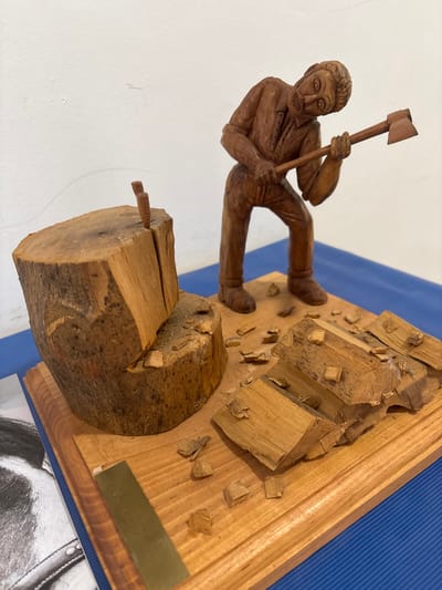 The image displays a detailed wooden diorama, located in El Espinar, Spain. The diorama rests on a polished, light-brown wooden base, which is situated on a blue corrugated surface. The central subject is a dark brown wooden figure of a bearded man, wearing a collared shirt and trousers, depicted in a dynamic pose. He is leaning forward, holding a small wooden axe with both hands, poised as if in the act of chopping. To the figure's left, a lighter brown wooden tree stump stands upright, featuring a prominent vertical split. A slender wooden wedge is inserted into the upper portion of this split. Several irregularly shaped pieces of lighter brown chopped wood and numerous smaller wooden splinters are scattered across the wooden base, predominantly to the figure's right and front. The background consists of a plain white wall. A partial black and white image, possibly a drawing or photograph of a horse's head with a bridle, is visible in the lower left periphery, underneath the wooden base.