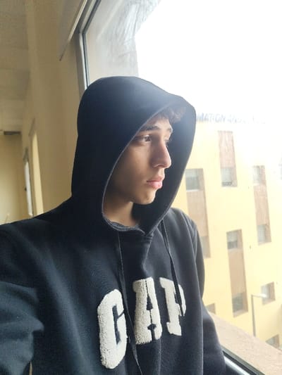 The image captures an individual in Jibb as-Safa, Syria, positioned indoors, looking out through a large window. The subject, appearing male with light skin and short, dark, curly hair, wears a black hooded sweatshirt with the hood covering the head. The front of the sweatshirt displays the word "GAP" in prominent, raised, white, textured letters. The individual is oriented in profile, facing towards the right side of the frame, gazing intently beyond the windowpane. The immediate interior background to the left of the subject features a plain, light beige or off-white wall, with a partial view of an arched opening further back. Through the window, an exterior scene is visible, dominated by a multi-story building with yellow-tan colored walls and numerous vertically aligned rectangular windows. Faint text, reading "FORMATION AN," is discernible on the upper portion of this exterior building. The exterior environment appears brightly lit and possibly overcast. A light fixture or pole is partially visible outside the window at a lower elevation.
