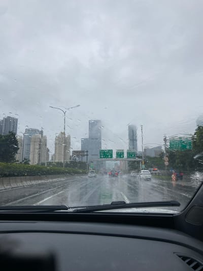 This image depicts a view from inside a vehicle driving on a highway in Luohu District, China, during a heavy rainstorm. The windshield is covered in raindrops, distorting the view of the cityscape ahead. Tall buildings, including a distinct skyscraper with a pointed top, are visible through the rain. Green highway signs with Chinese characters are present, indicating directions. Several cars are driving on the wet, reflective highway, their taillights visible. The sky is overcast and gray. The road appears slick and partially flooded in places. The interior of the car, including the dashboard and windshield wipers, is partially visible in the foreground.