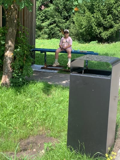 A young person wearing a pink t-shirt, shorts, sunglasses, and a cap sits on a blue park bench. The person's legs are crossed and they are holding a phone. The bench is situated in a grassy park area, with trees and foliage in the background. A black trash can is positioned in the foreground to the right of the bench. The scene appears to be in Arbuzov-Baran, Russia.