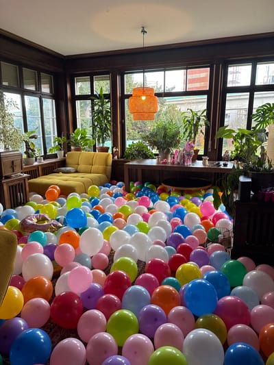 The image depicts an interior room located in Wallisellen, Switzerland, filled with a dense layer of multi-colored balloons covering the entire floor. The room features dark wood paneling and trim, a white ceiling, and large windows spanning at least two walls, providing natural light.
Furniture includes a mustard yellow, segmented sofa positioned in the left corner against the windows, a dark wooden cabinet on the far left, and a large, dark wooden table in the center-right, its surface partially visible above the balloons. An orange, tiered, woven pendant light fixture hangs from the ceiling.
Numerous potted green plants are arranged on the window sills and the large wooden table. On the table, a pink glitter unicorn figurine is prominently displayed alongside smaller pink decorative objects.
The balloons are inflated and appear to be latex, ranging in color to include pink, blue, green, yellow, orange, purple, white, and red. A large, metallic foil balloon shaped like a donut with sprinkles is visible among the other balloons in the lower-left foreground. No individuals are present in the scene. Through the windows, exterior greenery and parts of residential or commercial buildings are visible.