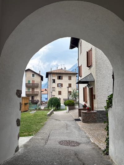 The image, taken in Molveno, Italy, depicts a view through a curved archway looking out onto a street scene. The perspective is from inside a building, looking outward, and the image is rotated 90 degrees counter-clockwise. The foreground is dominated by the textured white plaster of the archway.

Beyond the arch, a narrow street runs diagonally through the frame. On the right side of the street, there is a paved walkway with a circular metal grate embedded in the asphalt. A small patch of green grass borders the walkway. On the left side of the street, several buildings are visible. These appear to be residential structures with stone facades, wooden shutters on the windows, and balconies. Some of the buildings have flower boxes with red and white flowers. In the background, glimpses of mountains can be seen between the buildings, suggesting a mountainous or hilly region.

The sky above is partially cloudy, indicating daytime. There are no visible people in the scene, and the overall impression is one of a quiet, picturesque Italian town. The scene is well-lit, suggesting it was taken during daylight hours, likely mid-morning or afternoon. The texture of the buildings and the cobblestone walkway suggest an older, traditional European setting.