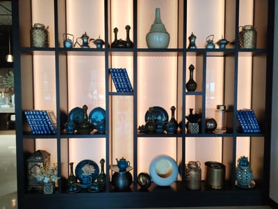 A dark wooden, multi-shelf display unit is filled with various decorative objects. The shelves are illuminated from behind with warm, soft light. On the top shelf, from left to right, are a gold woven vase, several silver teapots and covered bowls, and a light green ceramic vase with a long neck. The second shelf from the top features two dark green decorative vases, followed by a stack of blue-bound books, a collection of blue and green decorative plates and vases, and a tall, slender dark green vase. The third shelf holds a decorative lantern with dried flowers, a small blue teapot on a dark base, and several blue decorative plates and cups. The bottom shelf contains a gold woven lantern, a gold cylindrical container, and a light blue decorative lantern. The overall environment appears to be an indoor space, possibly a hotel lobby or a high-end retail store, with subtle hints of a dimly lit background and reflective surfaces. The location is identified as Konigil, Uzbekistan.
