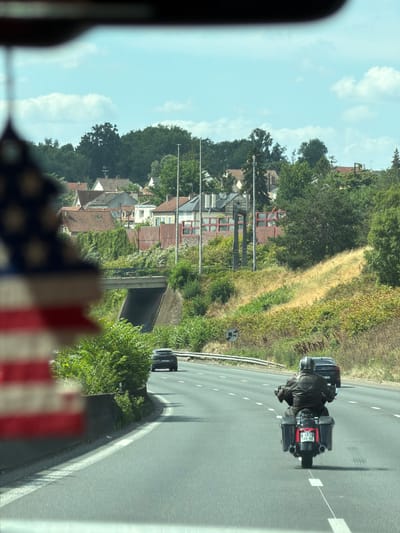The image shows a motorcycle rider from behind, traveling on a road in Savigny-sur-Orge, France. The rider is wearing a leather jacket and helmet, and the motorcycle is a touring model with saddlebags. The road appears to be a highway, with multiple lanes and white markings. The setting is rural, with trees and houses visible on the side of the road. The sky is blue with some clouds, suggesting it is daytime. The scene is captured from a slightly elevated perspective, possibly from another vehicle. A blurred American flag is visible in the foreground, obscuring a portion of the left side of the image. The license plate of the motorcycle has the text "AM 460 BX 69".