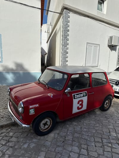 A red classic Mini Cooper with a white roof is parked on a cobblestone street in Costa da Caparica, Portugal. The car has a racing number "3" on its side with a Castrol sticker above it. There are also "MOTUL" and "SACOR" stickers on the front fender. The car is positioned at an angle, with its front facing left. To the right of the Mini, a white modern Mercedes-Benz is partially visible, showing its license plate, "NX 8-XX". The background consists of white buildings with visible architectural details such as windows and air conditioning units. The street is lined with cobblestones, and shadows are cast by the buildings and the car. The lighting suggests it is daytime, possibly late morning or early afternoon, with bright sunlight.