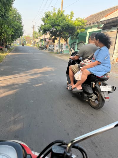 A couple rides a scooter down a street in Karangtanjung, Indonesia. The man is driving and the woman is riding behind him, holding a plastic bag. The street is lined with trees and buildings. The sky is clear and the sun is shining. The scooter's license plate is visible and reads "W 3472 NGB". The camera is positioned behind the couple, looking forward.