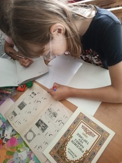 The image shows a young person with blonde hair, wearing glasses and a dark blue t-shirt with a floral print, leaning over a table and writing in a notebook. The person is holding a pen in their right hand and is focused on their work. There are several books and papers spread out on the table, some of which appear to contain text and illustrations in a foreign language, possibly Arabic or a related script. A red eraser and a green object, possibly a pencil sharpener, are also visible on the table. The environment suggests a study or learning setting, potentially at home or in a classroom. The image does not provide any specific visual cues to determine its exact location within the city of Buraly, Russia, or any other geographical context.