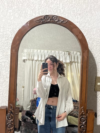 A mirror reflects a woman holding a phone, capturing her casual style. She wears a black top, jeans, and an unbuttoned white-striped shirt. Curtains hang behind her. The mirror has a dark wood frame with ornate detailing at the top and sides. The wall is a textured white. A light switch is visible above the mirror. Additional items like a rug, decorations, and furniture partially appear within the mirror's reflection.