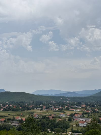 This is a wide, high-angle shot of a rural landscape in Vidrovan, Montenegro. The sky is filled with scattered white and gray clouds. In the middle ground, a village is nestled amidst green hills and fields. The village consists of numerous houses with red-tiled roofs, interspersed with trees and some open grassy areas. The houses vary in size and architectural style, with some appearing to be single-family dwellings and others larger structures. In the background, layered mountain ranges recede into the distance, their forms softened by the atmospheric haze. The overall impression is one of a peaceful, established rural community within a scenic natural environment.
