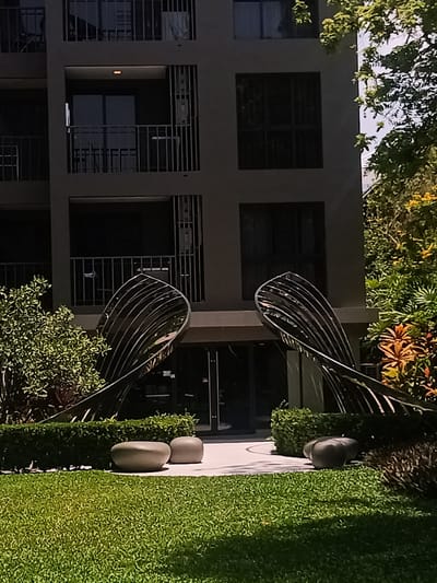 This photograph captures a scene in Hua Hin City Municipality, Thailand. The shot is taken at ground level, focusing on the entrance of a modern building. Two large, abstract, reflective metal sculptures, curved and lined, flank the entrance of the building. These sculptural elements rest on a raised concrete walkway.
In the foreground, a well-manicured lawn extends towards the viewer. Scattered across the walkway are several smooth, rounded stone or concrete seats, suggesting a place for visitors to pause and relax.
Behind the sculptures, the building is visible. It has multiple stories, with dark-framed windows and balconies. The overall color scheme of the building is a muted gray, contrasting with the green lawn and surrounding foliage.
To the right of the building, there is dense vegetation, including various trees and plants with orange and yellow flowers, indicating a tropical climate. The composition is balanced, with the architectural elements of the building offset by the natural greenery. The lighting appears soft and diffused, suggesting either an overcast day or a time of day when the sun is not directly overhead.