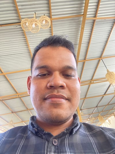 A man is shown from the chest up, looking directly at the camera in a selfie. He has dark hair and brown eyes, and is wearing a dark plaid shirt. The background is a corrugated metal ceiling supported by yellow beams. Several woven pendant light fixtures are visible hanging from the ceiling. The setting appears to be an indoor or covered outdoor space. There is no specific indication of location within Aluepiet, Indonesia.