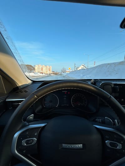 The image depicts a winter street scene in Dobryanka, Russia, viewed from inside a vehicle. The foreground is dominated by the black leather steering wheel of a Haval brand vehicle, featuring white stitching, paddle shifters, and various control buttons. The digital instrument cluster is visible through the steering wheel, displaying "HAVAL", a warning light, the time "17:36", and "1234 km". The dashboard is textured.

Outside the windshield, a clear, bright blue sky is visible above a snow-covered landscape. The road ahead, which appears cleared, is flanked by substantial snowbanks. In the mid-ground, a yellow diamond-shaped sign (indicating a priority road) is erected on the right side of the road. Further right, a small white building with a dark roof is partially obscured by snow, along with a street light pole and utility wires. In the distance to the left, a cluster of multi-story residential buildings, light-colored with visible windows and balconies, stands against the horizon. Bare deciduous trees are present on the left. The general environment is characterized by heavy snow cover and clear, cold weather. No individuals are visibly present in the scene.