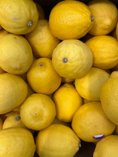The image is a close-up, overhead shot of a large pile of bright yellow lemons. The lemons are varied in size and shape, with some showing the characteristic bumpy texture of citrus fruit. A few lemons have small brown or blue spots on their skin, and some have visible stem ends. One lemon in the upper right has a small circular sticker with blue, white, and red segments, which appears to be a produce label. The lighting is bright, suggesting it might be daytime, and there are no discernible shadows indicating a specific time of day. The scene is likely a market stall or grocery store display, given the abundance and presentation of the fruit. There are no people visible in the frame. The image is a still photograph with no discernible activity taking place. The setting is not explicitly identifiable beyond being a place where lemons are stored or sold.