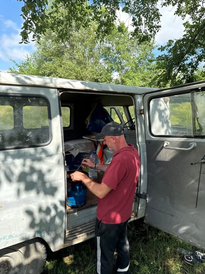 A middle-aged man wearing a maroon polo shirt and a black baseball cap is seen from the side, preparing food outdoors. He stands beside an open, light gray van with its rear door ajar. The man is actively engaged in cooking, holding a utensil over a small pan placed on a blue gas cylinder. A plastic bottle of water is also visible nearby. The scene appears to be set in a grassy area with trees and foliage in the background, suggesting an outdoor location. The light gray van has visible wear and tear, with some rust and peeling paint on its exterior. The overall environment indicates a casual, possibly camping or roadside setting. There is no information to confirm or deny the location being Khidiskuri, Georgia.