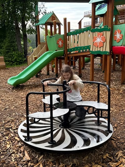 A young girl is playing on a spinning playground apparatus in a wood-chip-covered play area. The spinning apparatus is black metal with white and black patterned seats. Behind the girl is a green slide attached to a wooden playground structure with flower decorations. Trees and a building are visible in the background. This location is in the city of Peresvet, Russia.