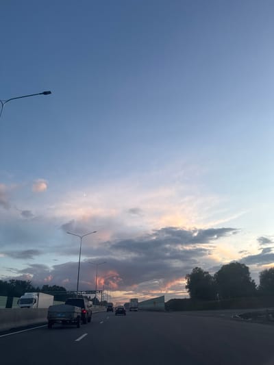 The image captures a twilight scene on a highway, likely near Dubki, Russia. The sky is a gradient of blue, with soft pink and orange hues illuminating the clouds, indicating the sun is setting or has recently risen. A crescent moon is visible in the upper portion of the sky.
Several vehicles are traveling on the multi-lane highway. In the foreground, a pickup truck pulling a trailer is in the right lane. To its left, a large white truck is in the left lane. Further down the highway, other vehicles, including cars and trucks, are visible. Streetlights line the side of the road, casting a faint glow.
On the left side of the highway, a sound barrier is present, with some vegetation visible beyond it. On the right side, trees and foliage line the embankment. The overall impression is one of a serene evening commute or journey on a highway.