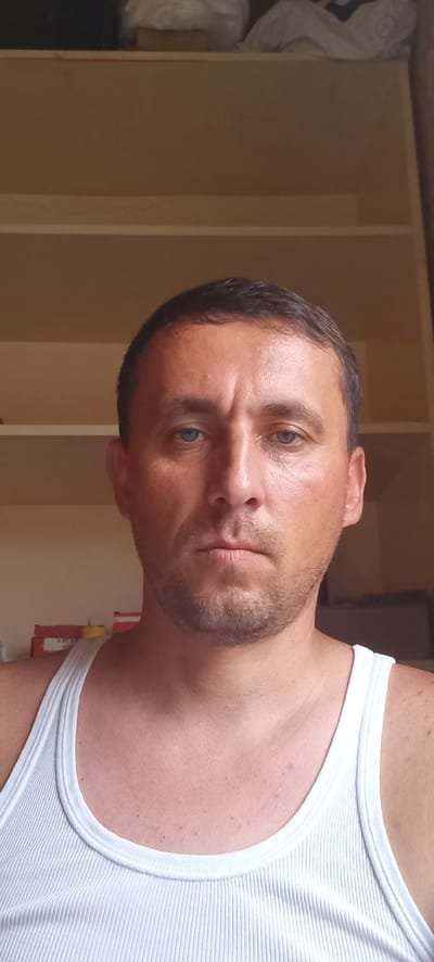 A medium shot shows a man with short dark hair, blue eyes, and light skin wearing a white ribbed tank top. He is looking directly at the camera with a neutral expression. He has a light stubble on his face and his skin appears tanned and slightly oily. The background consists of light brown shelves, with some out-of-focus items on them. The scene is indoors.