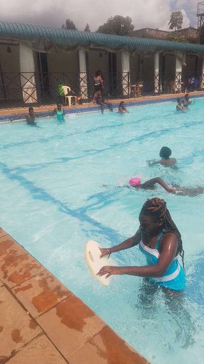 This image captures an active outdoor swimming pool scene located in Kikuyu Township ward, Kenya. The pool features clear, light blue water with lane lines visible on the bottom. The foreground shows terracotta-colored coping tiles bordering the pool.

Approximately ten individuals are present. In the immediate foreground, a female with braided hair, wearing a turquoise and white one-piece swimsuit, stands waist-deep in the water, holding a cream-colored kickboard with integrated handgrips. Further into the pool, one individual swims with their back to the camera, while another, wearing a pink swim cap, executes a freestyle stroke. To the left, a male is chest-deep in the water, and a female in a green swimsuit stands beside him.

Near the left edge of the pool, one person sits on the pool's edge with their legs submerged, supporting another individual who is perched on their shoulders. Along the far side of the pool, a building with a blue corrugated metal roof and several open, archway-like structures is visible. Inside these sheltered areas, multiple people are present, some standing, some seated. A white plastic chair with a green bag rests on the pool deck near the building. Green trees and distant structures are visible under a bright, partly cloudy sky beyond the pool facility.