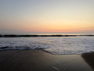 A serene beach scene unfolds under a soft, pastel-colored sky at sunset or sunrise. The sky transitions from light blue to hues of peach and gold. Gentle waves roll onto a dark, wet sand beach. The water is foamy and reflective. The horizon line is clear, with the ocean meeting the sky in a seamless blend. The overall mood is peaceful and tranquil.