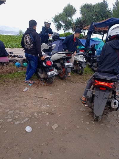 The scene shows a group of people gathered around several motorcycles parked on a dirt road at the beach in Pameungpeuk, Indonesia. The coordinates -7.668, 107.694 pinpoint the location to a beach area near the town of Pameungpeuk. There are four men and two motorcycles. The man in the white helmet is sitting on a white motorcycle, while another man is standing beside him.  A third man is sitting on a black motorcycle, and the fourth man is standing nearby. Several plastic water bottles and a plastic bag litter the ground. Green foliage and bushes are visible in the background. The sky is overcast with a cloudy sky. The group appears to be taking a break or getting ready to go.  The motorcycles are in good condition, with visible license plates.