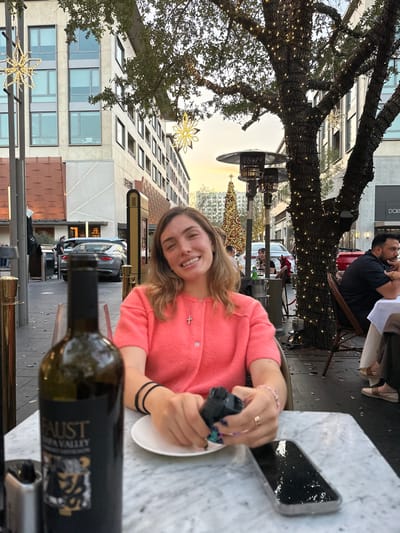 A woman smiles at the camera, seated at an outdoor table with a white marble top. She wears a pink sweater and a cross necklace. A bottle of wine labeled "FAUST" is in the foreground. In the background, a building with numerous windows and snowflakes adorns the tree branches. A Christmas tree stands further back on the street. Other patrons are seated nearby. A phone rests on the table.
