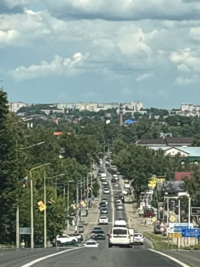 The image depicts a street scene in Pereslavl-Zalessky, Russia, viewed from a high vantage point, likely from a moving vehicle. The perspective is such that the road and buildings appear to be tilted. The scene is populated by numerous cars, indicating traffic. In the foreground, a white truck is prominent. Further back, rows of cars are visible on the road, suggesting a busy day. The surrounding environment is lush with trees, interspersed with apartment buildings of varying heights. The sky above is filled with white, fluffy clouds, suggesting fair weather. The presence of traffic lights and road signs indicates an urban or semi-urban setting. The overall impression is that of a typical day with moderate to heavy traffic in a Russian city. No discernible text is legible in the image.