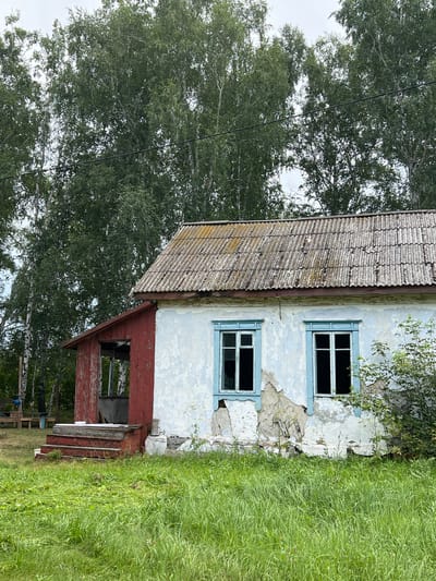An exterior, medium shot captures a weathered, abandoned house situated in the city of Курорт Озеро, Russia. The house, painted white with blue window frames, shows significant signs of decay, with peeling paint and crumbling plaster. A red-painted porch with wooden steps leads to an open doorway. Behind the house, a dense forest of birch trees forms a natural backdrop under an overcast sky. The foreground is covered in lush, green grass. No people are visible in the scene.