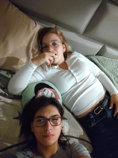 Two individuals are lying on a bed in what appears to be a bedroom. The person in the foreground is wearing glasses with maroon frames and a grey t-shirt. They have dark hair and a nose ring. The person in the background is wearing glasses with thin metal frames, a white long-sleeved shirt, and dark jeans with a black belt featuring a gold buckle. They have blonde hair. A decorative pillow with a cartoonish face design is positioned between the two individuals. The background includes a headboard of the bed and textured wall panels. The scene is indoors and the lighting suggests it is nighttime. The provided context indicates the location is Chorrillos, Peru.