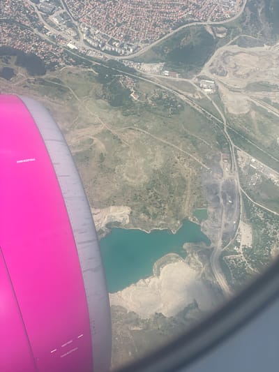 An aerial view from an airplane window shows a landscape with a body of turquoise water, a mine or quarry, and a residential area with red-tiled roofs. A portion of the airplane's pink engine cowling is visible in the foreground, with the text "COVER RECEPTACLE" and "HOIST POINT" printed on it. The residential area appears to be a dense collection of houses. The mine or quarry area features mounds of earth and what appears to be coal or dark material. Roads wind through the landscape, connecting different sections. The overall scene depicts a somewhat industrial or developed area adjacent to a natural landscape.
