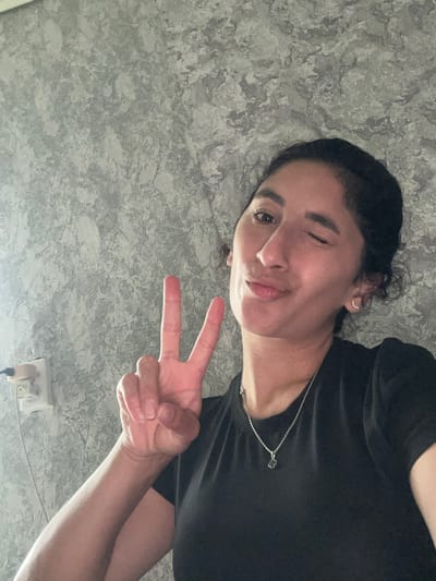 A young woman with dark hair and brown eyes is pictured in a selfie, posing against a textured grey wall. She is wearing a black t-shirt and a silver necklace with a small pendant. She winks and holds up two fingers in a peace sign with her right hand. An electrical outlet with a white cord is visible on the wall to her left. The image does not contain any specific details that would allow for the determination of its location within the city of Dondukovskaya, Russia.