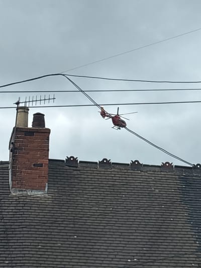 A red toy helicopter is suspended by its skids on a power line, its tail rotor appearing to be snagged on a higher wire. The helicopter is positioned to the right of a brick chimney topped with two vents and an aerial. In the foreground, a dark, tiled roof with decorative elements along its edge spans the width of the image. The sky is overcast and grey. The image was taken in Stoke-on-Trent, United Kingdom.
