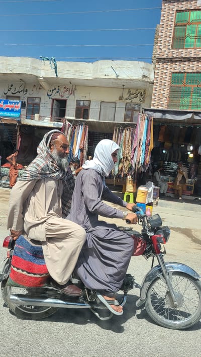 This image captures a vibrant street scene in Kuchlak, Pakistan, under a clear, bright blue sky. Two men are prominently featured on a red and black motorcycle with chrome accents, which appears to be stopped or moving slowly. The driver, wearing a white head covering and a striped shalwar kameez-style shirt, holds the handlebars with a watch visible on his left wrist. The passenger, an older man with a long white beard, is seated behind him, draped in a plaid scarf or shawl over his light-colored traditional attire. A colorful striped mat is placed on the motorcycle's rear seat.

The background reveals a bustling commercial street lined with multi-story buildings, likely shops. Rolls of colorful fabrics and textiles hang abundantly outside what appears to be a textile or clothing store, stretching along the sidewalk, suggesting a vibrant market. Other individuals can be seen inside the shops, adding to the sense of activity. The architecture includes buildings with exposed brickwork on upper floors and simpler, painted lower sections with visible windows, some featuring green-tinted glass.

The bright daylight is evident from the strong shadows cast on the asphalt road surface and the clear blue sky overhead, crisscrossed by utility lines. Visible text on shop signs includes "ار ساز کمالی" (Ar Saaz Kamali) and "حتی ارمین عالی واسکٹ محل" (Hatta Armeen Aali Waistcoat Mahal), along with several phone numbers (05438172426, 030563742, 0305-3591769), confirming the local context and commercial nature of the area. The scene captures a typical moment of daily life and commerce in a lively market setting in Kuchlak, Pakistan.